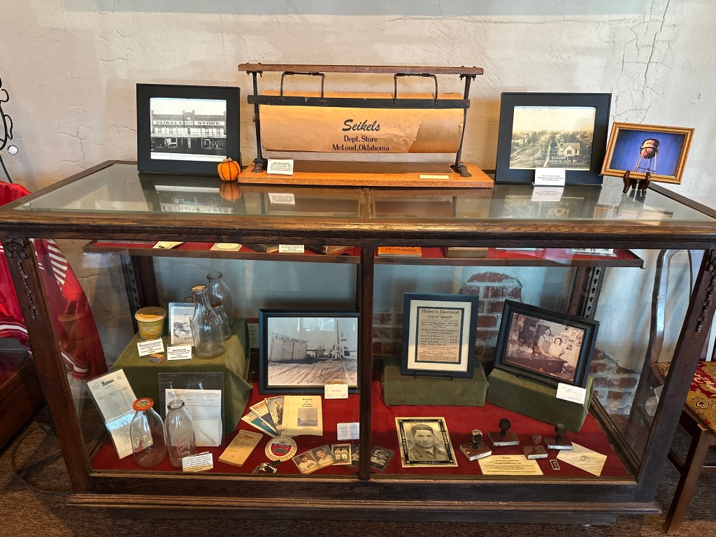 A display case inside the McLoud Historical Society Museum featuring various historical items, including photographs, documents, and memorabilia from local businesses.