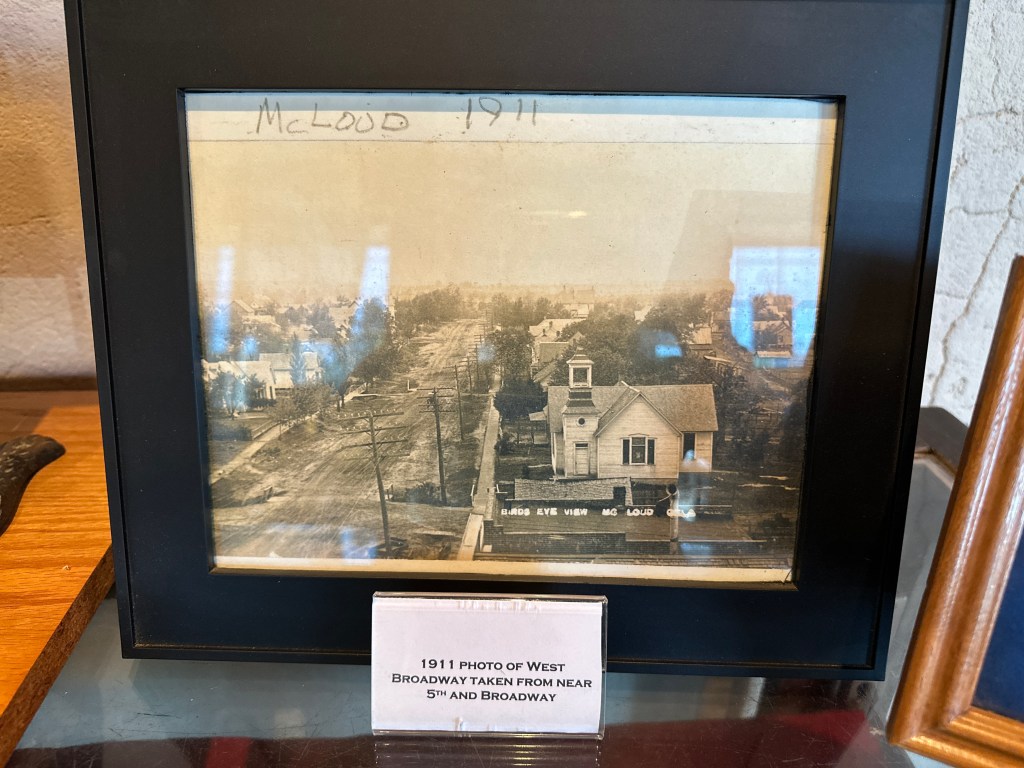 A framed black and white historical photograph labeled 'McLoud 1911', depicting a view of West Broadway taken from near 5th and Broadway, showcasing buildings and a church in McLoud.