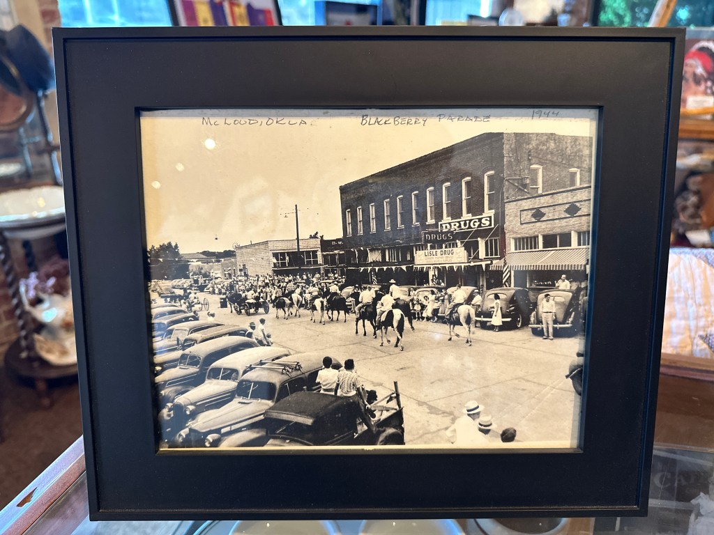 Historical photograph of a Blackberry Festival parade in McLoud, Oklahoma in 1944, featuring horses and a line of vintage cars in front of a drugstore.