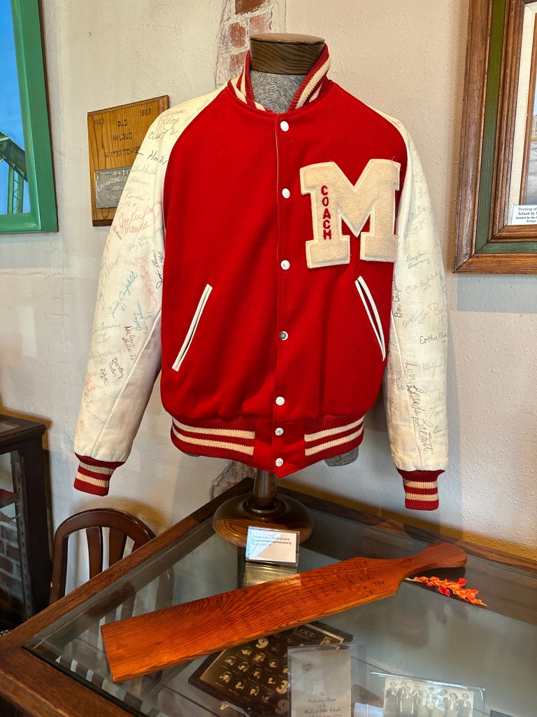 A red and white letterman jacket with the letter 'M' and 'COACH' embroidered on it, displayed on a mannequin in a museum, alongside a wooden paddle and other historical items.