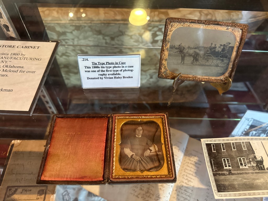 A display case featuring a tin type photo case from the 1800s, with one photo showing a seated individual and another photo depicting a horse-drawn carriage.