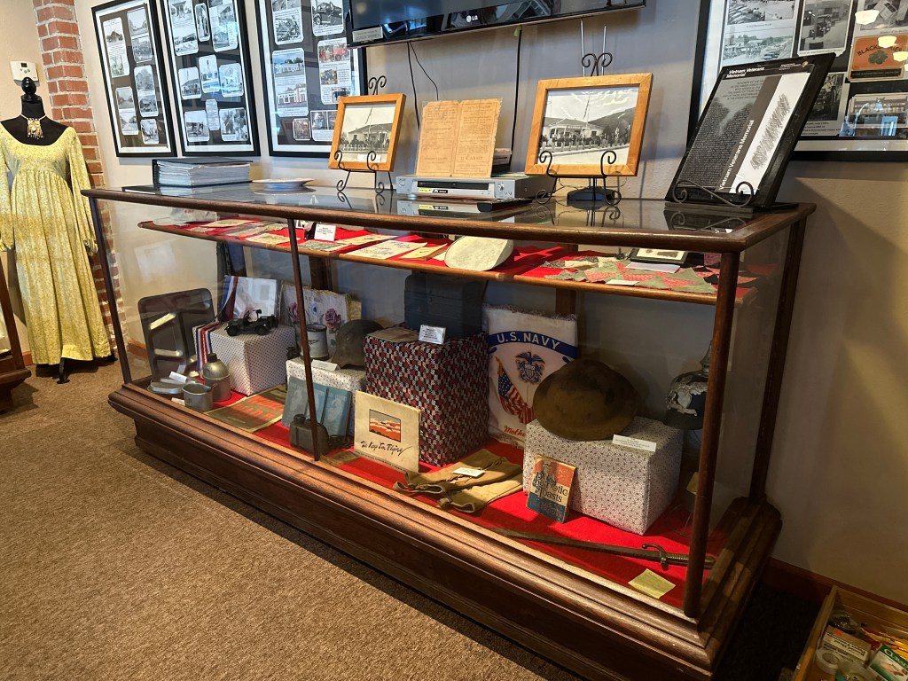Display case showcasing various historical artifacts and documents, including military items, photographs, and a vintage dress, set inside the McLoud Historical Society Museum.