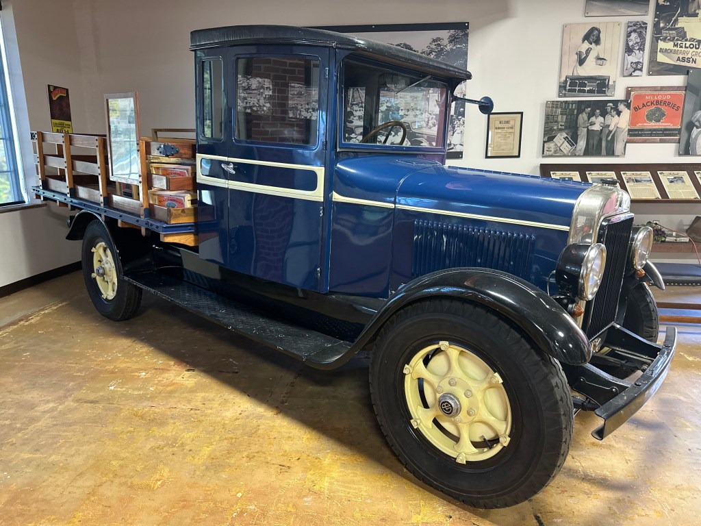 A vintage blue truck with a wooden flatbed, displayed inside the McLoud Historical Society Museum.