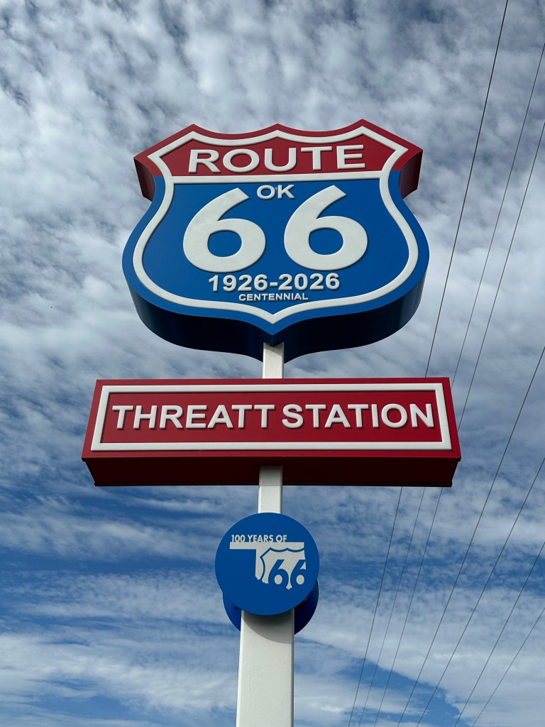 A sign for the Threatt Filling Station along Route 66, featuring the Route 66 shield and a 100-year anniversary emblem against a cloudy sky.