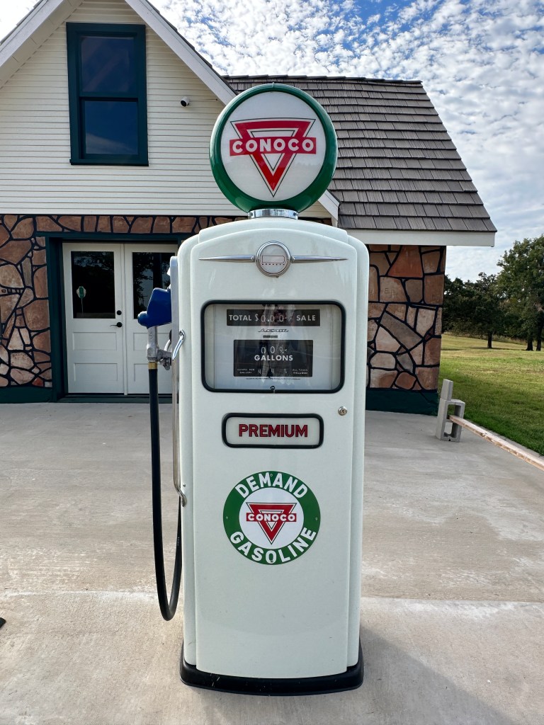 A vintage Conoco gasoline pump in front of the Threatt Filling Station, showcasing the building's unique architecture and green landscape under a partly cloudy sky.