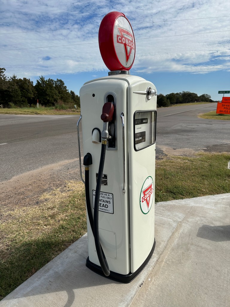A vintage Conoco gas pump with a rounded red top and signage displaying the Conoco logo, positioned beside a rural road under a partly cloudy sky.