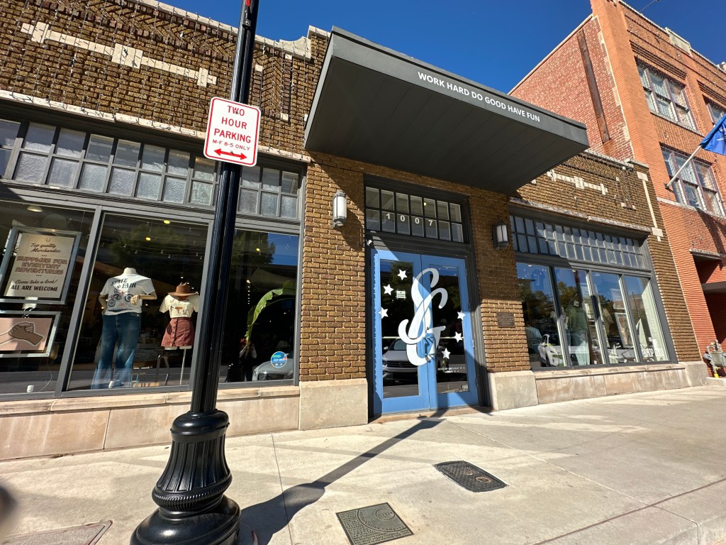 A storefront located on Automobile Alley in Oklahoma City, featuring a blue door with decorative elements and display windows showcasing clothing items. Above the entrance, a sign reads 'WORK HARD DO GOOD HAVE FUN.'