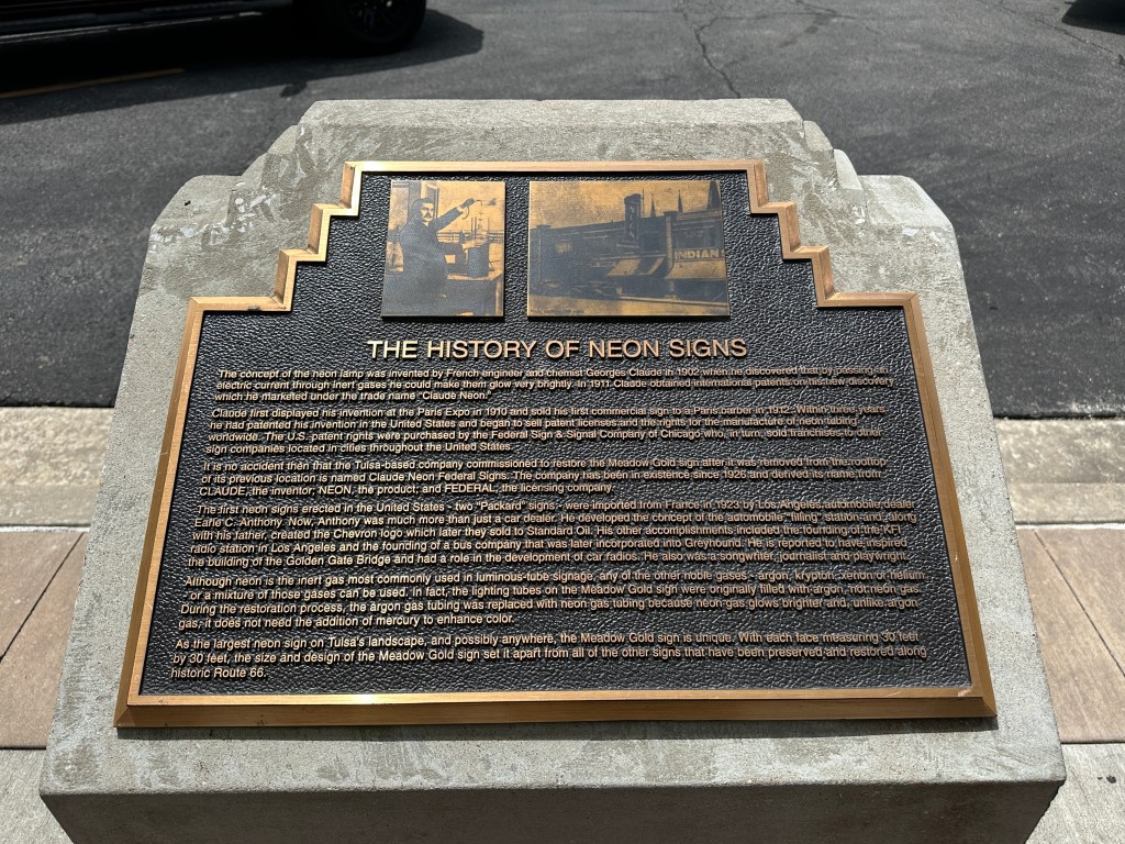 A historic plaque detailing the invention and history of neon signs, featuring engraved text and images, located near the preserved Meadow Gold sign in Tulsa, Oklahoma.