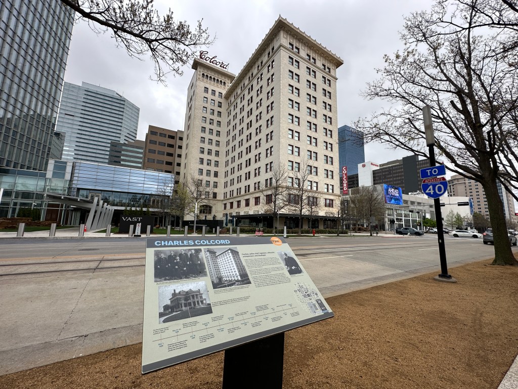 View of a historic building with a sign about Charles Colcord in the foreground, surrounded by modern skyscrapers and a street leading to Interstate 40.