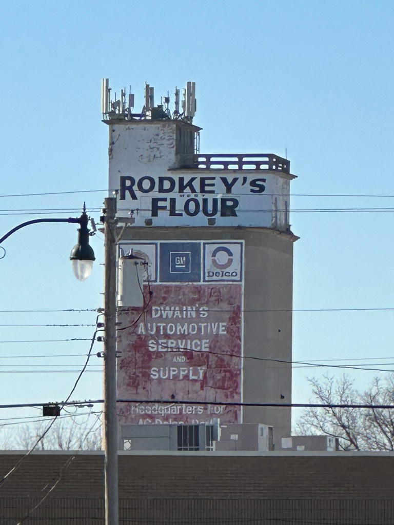 View of the Rodkey Flour Mill, featuring the iconic sign that reads 'Rodkey's Flour,' alongside Dwain's Automotive Service and Supply, set against a clear blue sky.