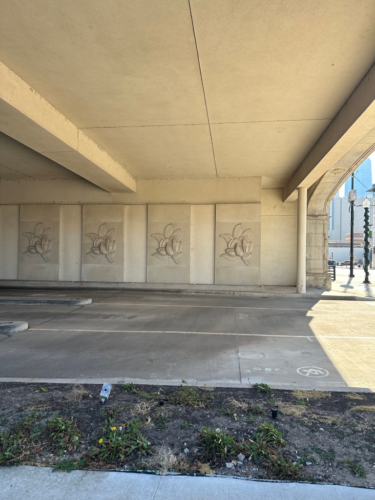 View of the underside of the Magnolia Petroleum Building, showcasing decorative floral reliefs on the wall, with a concrete structure overhead and parking space below.
