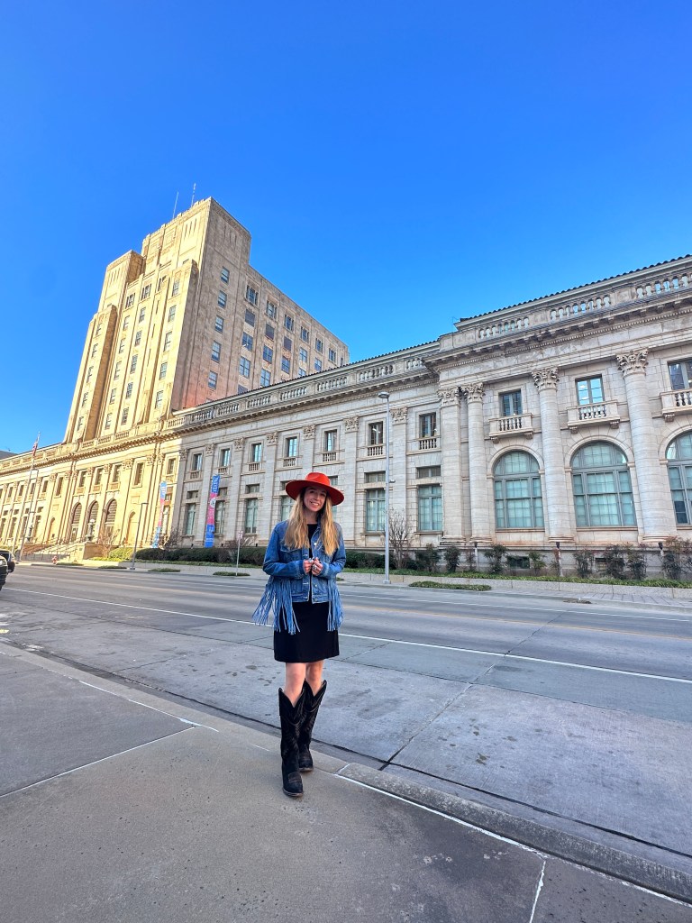 A woman stands on the street in front of a historic building, wearing a fringed denim jacket, a black dress, and cowboy boots, with a wide-brimmed red hat, against a clear blue sky.
