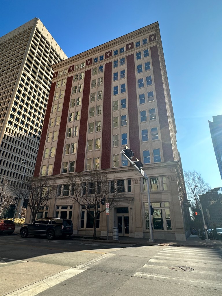 Historic brick building with large windows, located on a city street next to a modern structure, under a clear blue sky.
