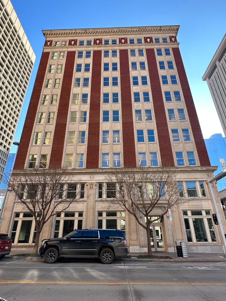 An ornate brick building with multiple windows, featuring a combination of red bricks and light-colored stone, situated on a city street. Two trees are in front of the building, and a large black vehicle is parked along the curb.