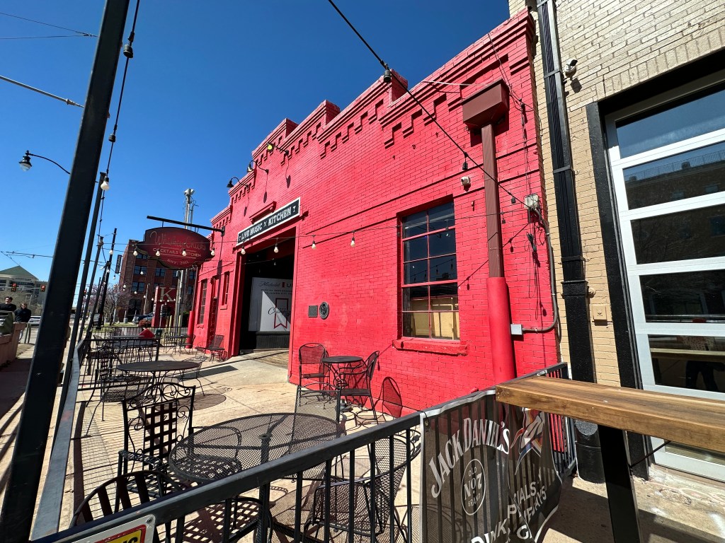 Exterior view of a vibrant red building with outdoor seating area, featuring tables and chairs, under a clear blue sky.