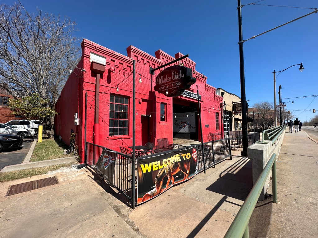 A bright red building with a sign for 'Whiskey Chicks' featuring an outdoor seating area, located along a sidewalk with trees and streetlamps in the background.
