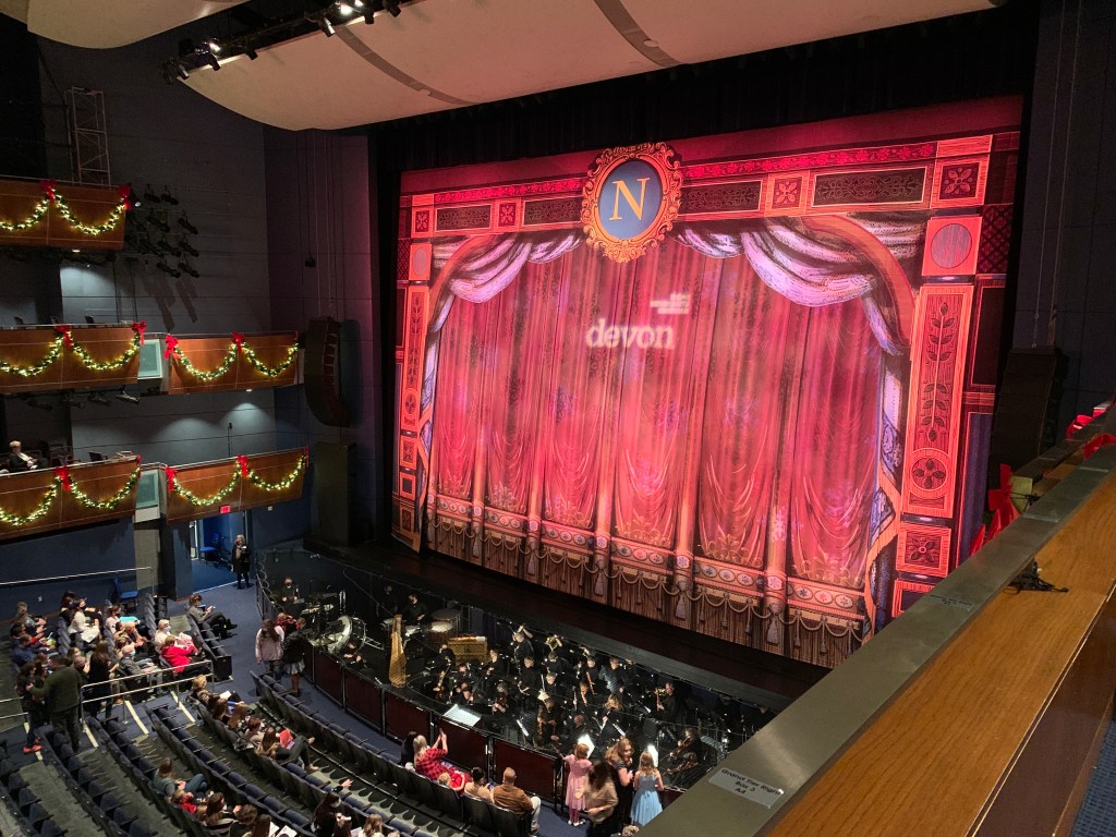 A view of a theater stage with a rich, red curtain and ornate decorations. The audience is seated in a dimly lit auditorium, with holiday decorations visible in the background.