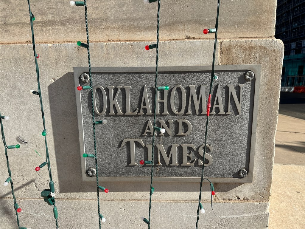 A brass plaque reading 'Oklahoma and Times' attached to a stone wall, surrounded by decorative holiday lights.