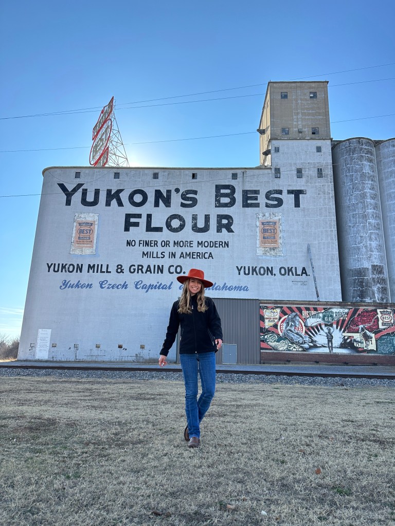 A woman in a red hat walks in front of a large building wall advertising 'Yukon's Best Flour' with the text 'No finer or more modern mills in America' in Yukon, Oklahoma.