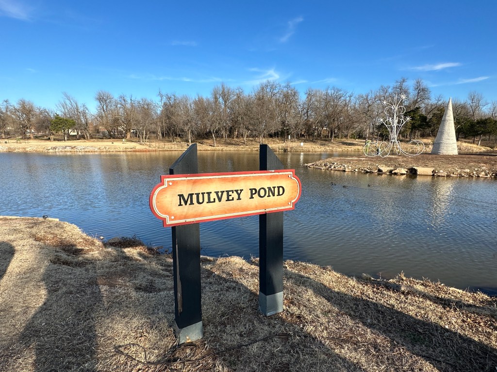 Sign for Mulvey Pond next to a serene pond surrounded by trees and a stone sculpture in the background.