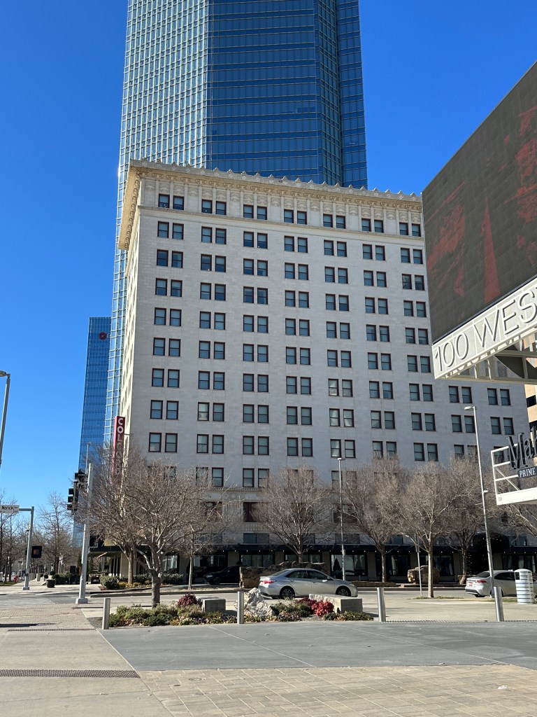 A historic low-rise building with numerous windows, located at the forefront of a modern high-rise skyscraper, under a clear blue sky.