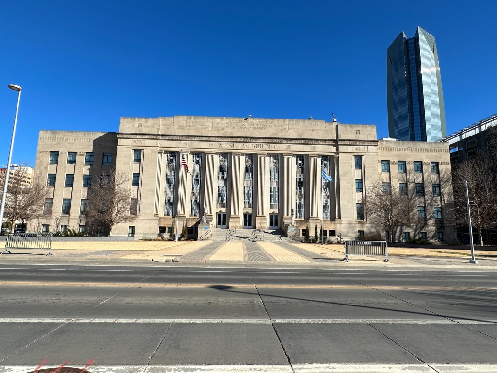 Front view of a municipal building with a prominent entrance, flanked by trees and flags, set against a clear blue sky, next to a modern skyscraper.