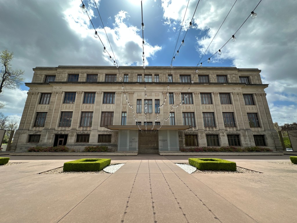 A historic building with a detailed facade, featuring large windows and decorative elements, set against a cloudy sky. Outdoor gardens with well-maintained hedges and light bulbs strung above.