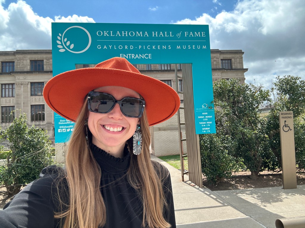 A smiling woman wearing a large orange hat and sunglasses stands in front of the Oklahoma Hall of Fame Gaylord-Pickens Museum entrance sign on a sunny day.