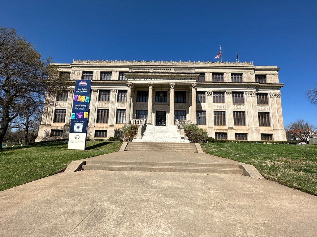 View of a large historical building with a grand entrance and stairs, surrounded by greenery and blue sky.