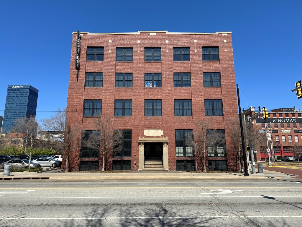 A four-story brick building with large windows and a central entrance, showcasing a modern urban architectural style against a clear blue sky.
