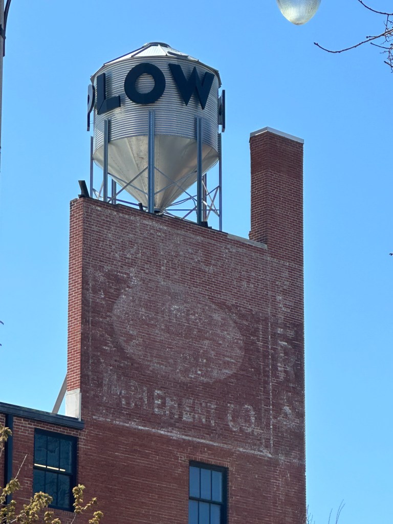 A close-up view of a brick building featuring a water tower with the word 'FLOW' on it, and an old faded advertisement for 'Ingredient Co.' on the side.