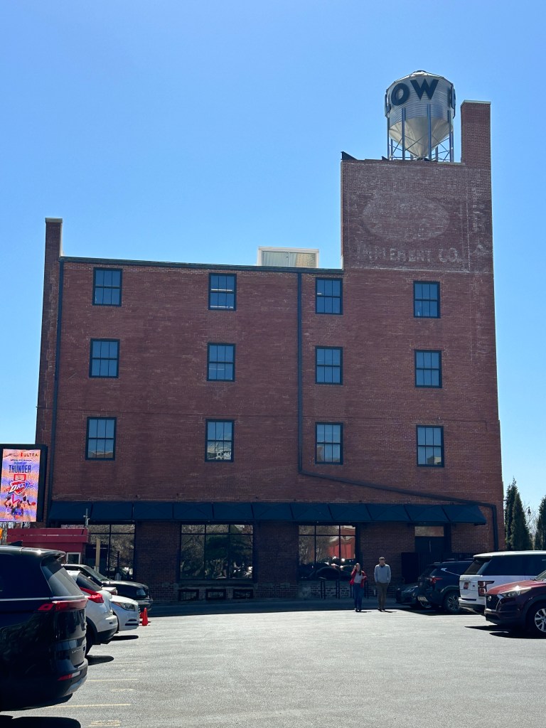 Exterior view of a red brick building with several windows and a water tower on top, surrounded by parking spaces and some people walking nearby.