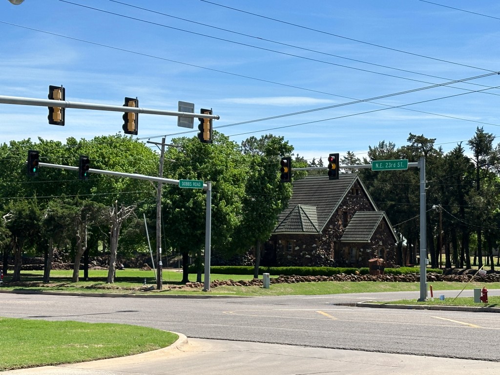 Street intersection with traffic lights and directional signs, featuring a stone house and green trees in the background.