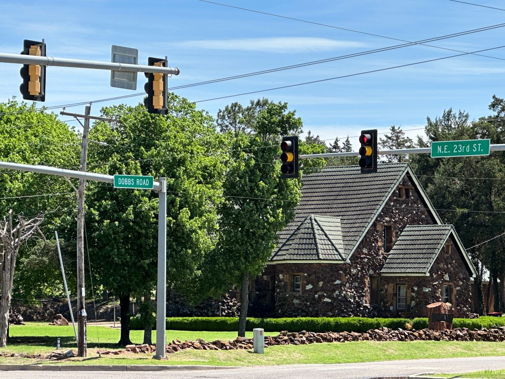 Traffic lights and street signs at the intersection of Dobbs Road and N.E. 23rd St. near a stone house surrounded by greenery.