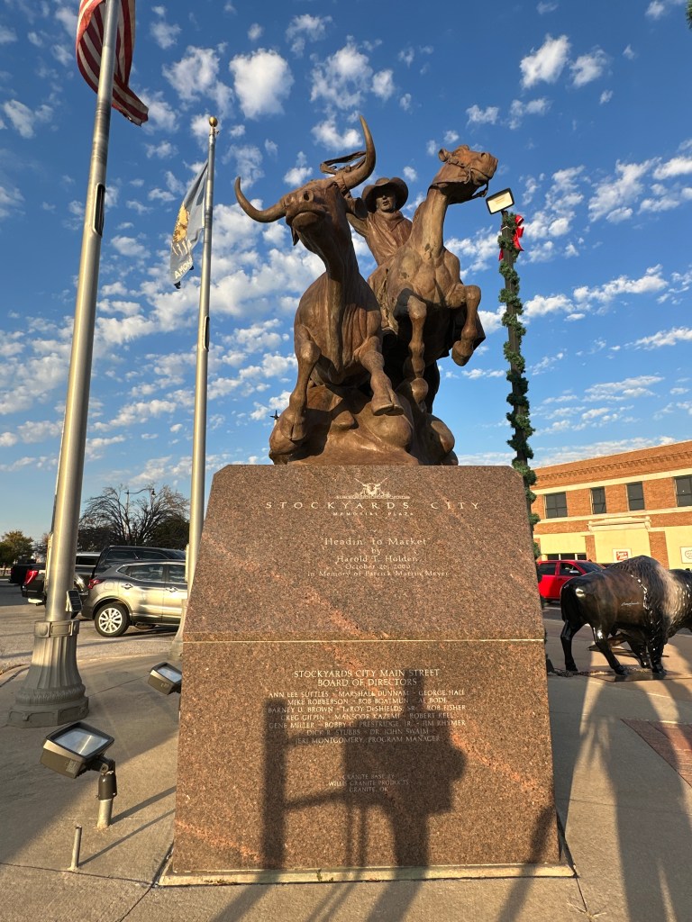 Bronze sculpture of a cowboy on horseback, guiding two cattle, with a commemorative plaque in front, set against a blue sky with clouds.