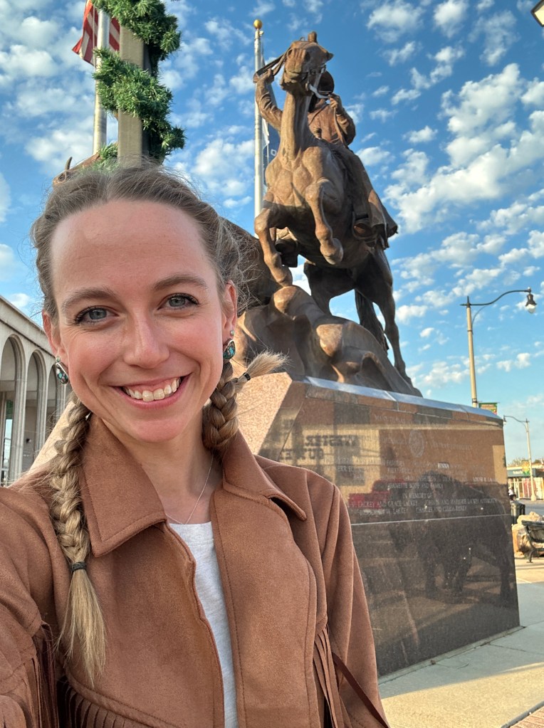A smiling woman with braided hair poses for a selfie in front of a large equestrian statue against a blue sky with clouds. The statue depicts a mounted figure holding a flag, located near a street with decorative elements.
